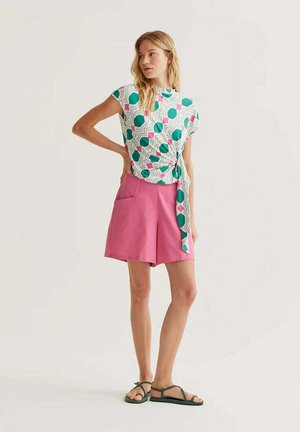 Mujer de pie con la mano en la cadera, lleva una blusa blanca con estampado de formas verdes y rosas, shorts rosas de talle alto y sandalias.