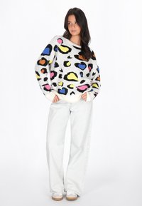 Pull en tissu doux, blanc avec des formes abstraites multicolores vives. Présente une coupe décontractée et des poignets côtelés. Associé à un pantalon en denim clair.