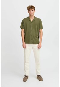 Camisa de manga corta en verde oliva con cuello, tapeta de botones y textura suave, combinada con pantalones blancos y zapatos de gamuza marrón.