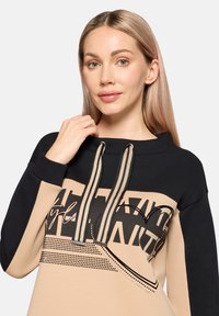 Sweat à capuche noir et beige, arborant des graphismes audacieux et des motifs à pois. Cordons de serrage contrastants et coupe décontractée.