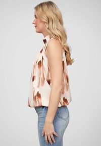 Débardeur sans manches en tissu rose clair avec des motifs abstraits marron. Présente un col haut et une coupe loose, associé à un jean délavé.