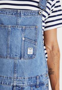 Des salopettes en denim bleu clair avec des bretelles réglables, une poche avant et une étiquette cousue. Un t-shirt ray�é est visible en dessous.