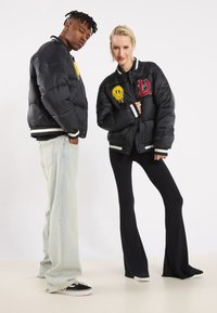Black puffer jackets with embroidered yellow smiley and red lettering, paired with light blue jeans and black pants. Features striped cuffs.