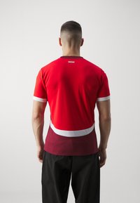 Rød atletisk t-shirt med rund hals, korte ærmer og en hvid accentstribe. Stoffet ser glat og letvægtsagtigt ud.