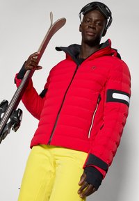 Rote Pufferjacke mit Reißverschluss, schwarzen Akzenten und weißen Streifen. Kombiniert mit gelben Skihosen. Hält Skier und trägt eine Skibrille.