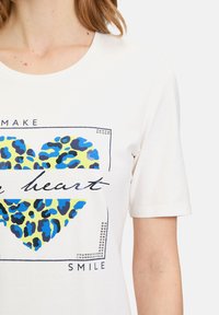 T-shirt en coton blanc avec un design de cœur en imprimé léopard bleu et jaune, accompagné du texte noir "MAKE YOU SMILE" et de clous décoratifs.