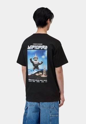 Persona che indossa una maglietta nera con una grande grafica di una figura mostruosa e uno skyline urbano sulla schiena, abbinata a pantaloncini cargo in denim blu.