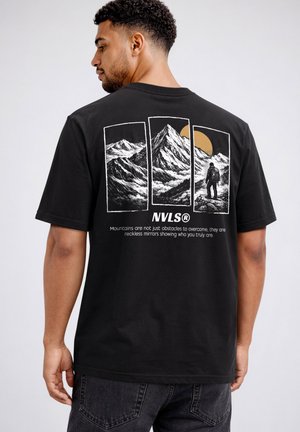Mann trägt schwarzes T-Shirt mit dreiteiliger Grafik einer Berglandschaft, Wanderer und gelber Sonne auf dem Rücken.