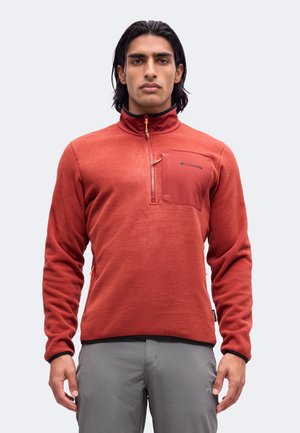 Άνδρας που φοράει fleece Columbia σε σκουριασμένο χρώμα με ημί-φερμουάρ και τσέπη στο στήθος, στέκεται μπροστά από ένα απλό φωτεινό φόντο.