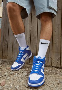 Las zapatillas Nike de caña alta presentan un diseño de cuero blanco y azul con una caja de los dedos perforada, suelas blancas y cordones azules. Se llevan con calcetines blancos de Nike.