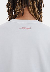 Lys grå sweatshirt med rund halsudskæring, der har rød broderet "Ed Hardy" branding på ryggen, hvilket fremhæver et blødt, tekstureret stof.