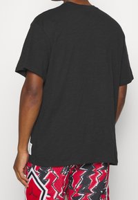 Persona con una maglietta nera a maniche corte semplice e pantaloni corti a zigzag rosso, nero e bianco, di spalle rispetto alla fotocamera.