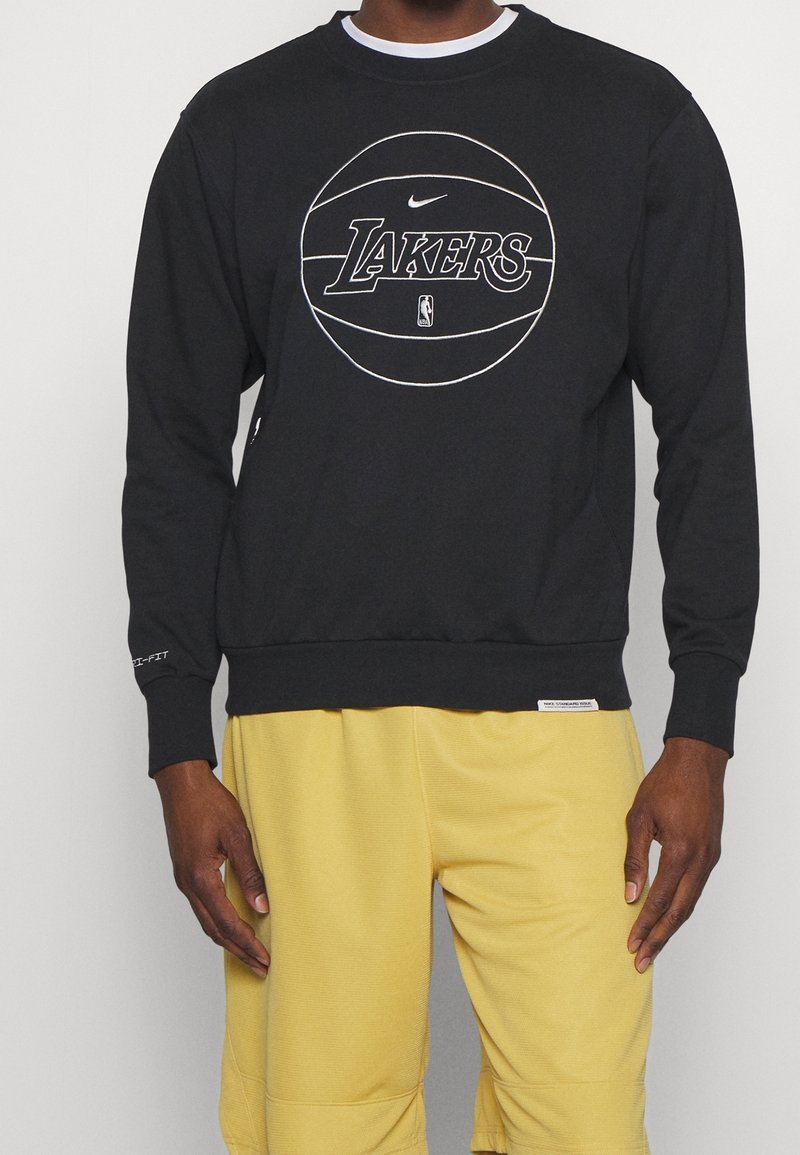Sudadera negra de Nike con un gráfico de baloncesto y el texto "LAKERS". Puños y dobladillo acanalados, textura suave, llevada con pantalones cortos amarillos.