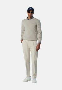 Maglione beige chiaro, pantaloni bianchi plissettati, berretto blu navy, occhiali da sole scuri e scarpe da ginnastica bianche. L'outfit presenta una camicia a quadri sotto.