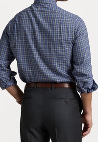 Man som bär en blå rutigt långärmad skjorta instoppad i mörkgråa kostymbyxor med ett brunt läderbälte, vänder bort med händerna vid sidorna.