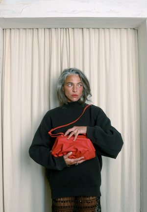 Sac en cuir rouge avec une forme courbée et une fermeture éclair, porté par une silhouette vêtue d'un sweat noir devant un fond de rideau clair.