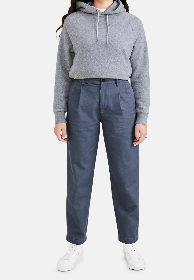 Grå kortärmad hoodie med snörning, kombinerad med mörkblå, högmidjade plisserade byxor och vita sneakers. Platt fram, avslappnad passform.