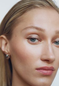 Gros plan sur le visage d'une personne montrant une texture de peau lisse, des lèvres rose pâle et des yeux bleu-vert. Les cheveux sont raides et blonds, accompagnés de boucles d'oreilles en argent simples.
