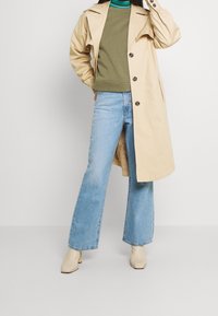 Abrigo trench beige con grandes botones, combinado con una sudadera verde oliva y vaqueros acampanados azul claro. Botines crema completan el conjunto.