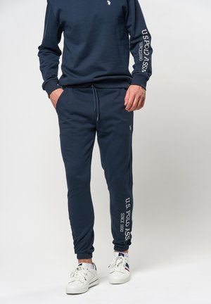 Man in marineblauwe U.S. Polo Assn. trainingsbroek en bijpassend sweatshirt met witte sneakers, staand met handen in de zakken.