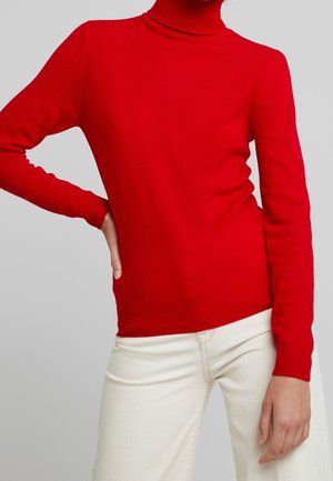 Persona con un suéter ajustado de cuello alto rojo y pantalones blanco hueso, con una mano en la cadera y la otra relajada a su lado.