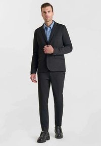 Completo grigio scuro dal taglio su misura, con blazer monopetto e pantaloni abbinati. Indossato sopra una camicia azzurro chiaro con colletto. Scarpe nere.
