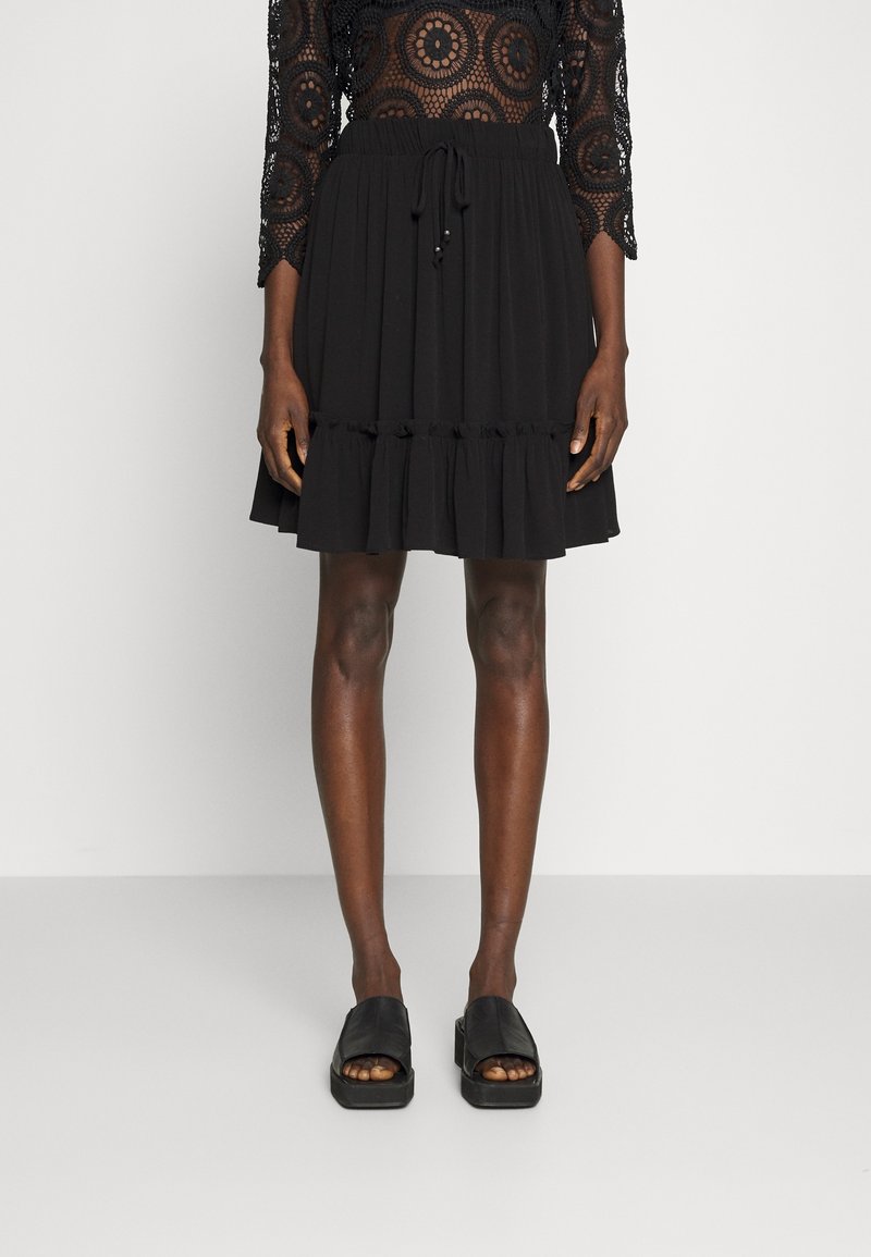 Jupe noire longueur genoux avec ourlet à volants, associée à des sandales à plateforme noires à bout ouvert, portée par une personne avec un haut en dentelle noire.
