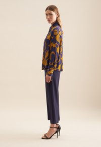Langsleeve blouse in paars en mosterd abstract patroon, gecombineerd met marineblauwe high-waisted broek en zwarte strappy hoge hakken.