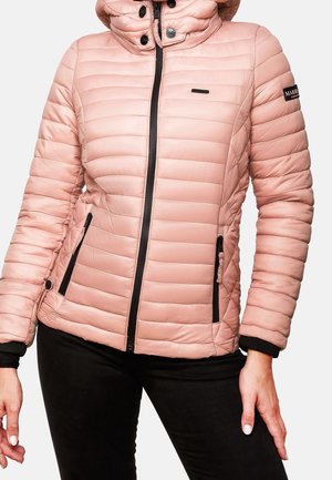 Woman wearing a light pink quilted jacket with black zipper details and black pants, standing with one hand on hip and one relaxed.