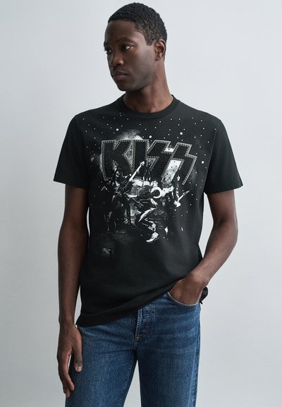 Man wearing black KISS band t-shirt with silver studded logo and graphic, paired with blue jeans, standing against plain light background.
