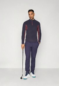 Navy modra flis majica z bordo poudarki, z zadrgo na ovratniku, v kombinaciji z navy hlačami in belimi golf čevlji, drži golf palico.