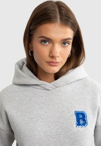 Šedá mikina s texturovanou tkaninou, na levé straně hrudi s vyšitým modrým písmenem "B" a slovem "Boosted" v bílém písmu.