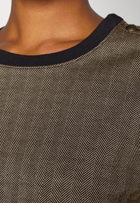 Brun och beige herringbone-mönstrad tyg med en svart ribbad krage och dekorativ knappdetalj på axeln.