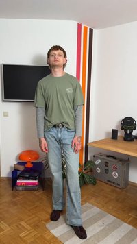 Πράσινο T-shirt φορεμένο πάνω από γκρίζο μακρυμάνικο πουκάμισο, συνδυασμένο με ανοιχτό μπλε τζιν και σκούρα παπούτσια. Φέρει τατουάζ και μαύρη ζώνη.