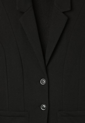 Blazer nero con revers a punta, dotato di chiusura a due bottoni, tessuto strutturato e due tasche frontali. Dettagli delle cuciture verticali visibili.