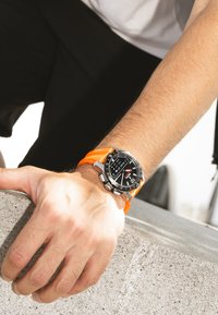 Svart digital sportklocka med orange silikonrem, silverdetaljer, har en rund urtavla och olika klockfunktioner visade.