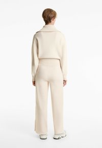 Pull en tricot beige et pantalon large assorti, tous deux avec une texture douce. Le pull possède un col ouvert et des poignets côtelés.