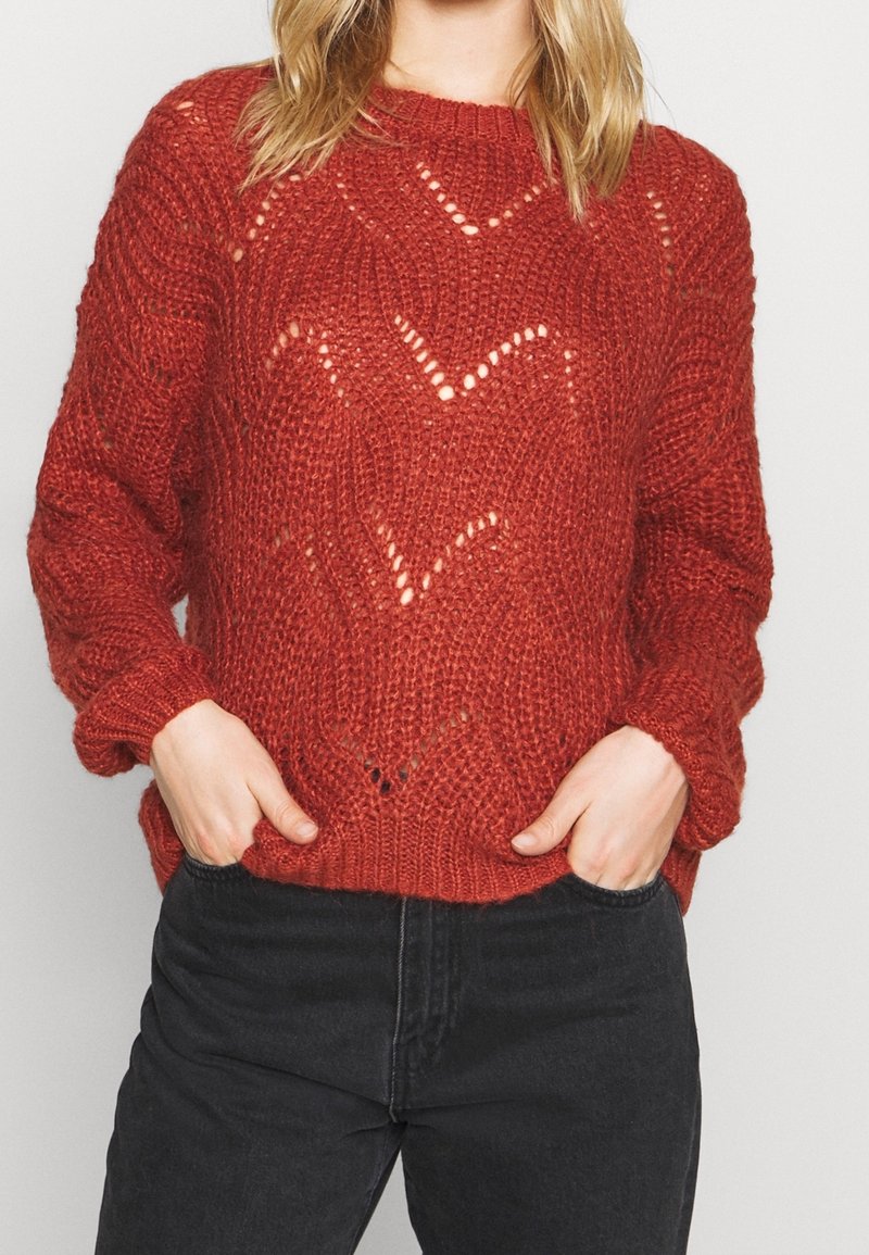 Personne portant un pull rouge tricoté avec un motif chevron et un pantalon noir, les mains dans les poches avant.