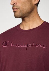 Mann in einem kastanienbraunen T-Shirt mit gesticktem "Champion"-Logo auf der Brust, vom Hals bis zur oberen Körperhälfte gezeigt.