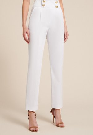 White high-waist tailored pants with gold buttons worn by a standing person wearing brown strappy high-heeled sandals on a neutral background.