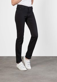 Persoon draagt strakke zwarte jeans en witte sneakers met klittenbandsluitingen, staande op een grijze betonnen vloer tegen een effen witte achtergrond.