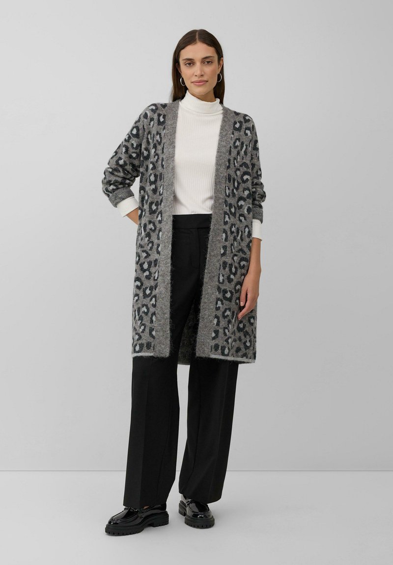 Femme portant un long cardigan à imprimé léopard gris, un col roulé blanc, un pantalon large noir et des mocassins noirs, se tenant devant un fond gris.