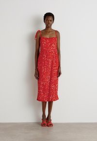Robe midi en sequins rouges avec des bretelles spaghetti, un buste ajusté et une jupe évasée. Présente des accents sur les bretelles et une texture lisse. Portée avec des sandales rouges.