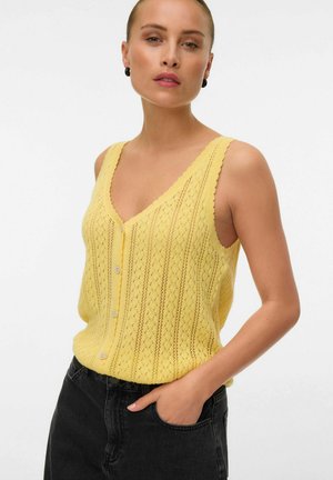 Femme portant un haut en tricot jaune sans manches à boutons avec un col en V, assorti à un jean noir, posant avec une main dans la poche.