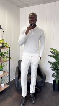Witte gebreide trui met een halve rits kraag, gecombineerd met op maat gemaakte witte broek en zwarte instappers. Binnenomgeving met planten en planken.