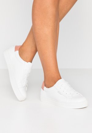 Jambes portant des baskets blanches basses avec des accents roses au talon, debout sur une surface lisse gris clair avec un fond assorti.