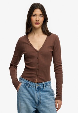 Jeune femme aux longs cheveux bruns portant un cardigan marron ajusté boutonné et un jean en denim bleu clair taille haute, les mains dans les poches.