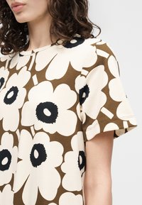 Γυναίκα που φοράει καφέ και κρεμ κοντομάνικο τοπ με μεγάλα λευκά λουλούδια με μαύρο κέντρο, με μπούκλες μαλλιών ορατές στους ώμους.