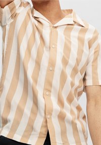 Camicia a maniche corte a righe beige e bianche, con colletto a punte e chiusura frontale con bottoni. Realizzata in tessuto leggero.