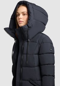 Jachetă puf din material bleu marin, cu o glugă mare, design matlasat, fermoar frontal și șnururi ajustabile. Prezintă un logo pe mâneca stângă.